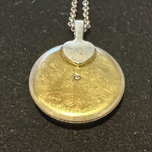May include: A round, gold-toned pendant with a small diamond accent. The pendant has a silver-colored bail and a heart-shaped detail. It hangs from a delicate silver chain, creating a classic jewelry piece.