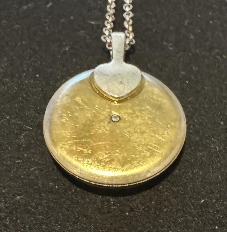 May include: A round, gold-toned pendant with a small diamond accent. The pendant has a silver-colored bail and a heart-shaped detail. It hangs from a delicate silver chain, creating a classic jewelry piece.