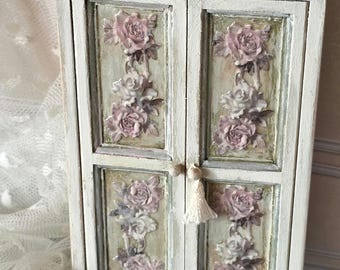 May include: A small, off-white wooden cabinet with two doors featuring raised, dusty pink rose floral reliefs on a muted green background. The cabinet has a rectangular top, a base with small feet, and a single white tassel pull on the right door.
