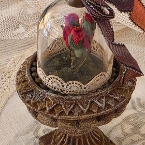 May include: A decorative cloche with red roses inside, set on a detailed, aged-looking pedestal. The glass dome is trimmed with lace, and a decorative ribbon is attached. The pedestal is brown with carved details, creating a vintage aesthetic.