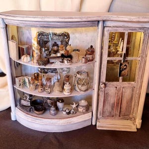 Puede incluir: Una vitrina en miniatura con frente de cristal curvo y tres estantes llenos de botellas diminutas, libros y objetos decorativos, junto a una puerta de madera con panel de cristal. La estructura tiene un acabado blanco desgastado.