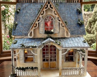 Peut inclure: Une maison de poupée miniature dotée d'un toit en bardeaux bleu vieilli, de murs couleur crème et de boiseries complexes. La structure comprend un porche avec une clôture blanche, des fenêtres arquées et de la verdure grimpante.