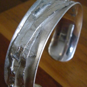 May include: Silver cuff bracelet with a textured, layered design. The bracelet is made of polished metal and has a smooth, rounded edge.