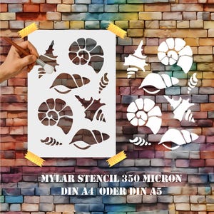Könnte beinhalten: Eine weiße Mylar-Schablone mit verschiedenen Muschelmotiven, darunter Spiralen und Fächerformen. Die Schablone ist mit gelbem Klebeband an einer Backsteinmauer befestigt. Der Text "MYLAR STENCIL 350 MICRON DIN A4 ODER DIN A5" ist aufgedruckt.