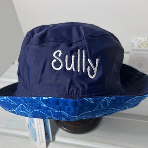 May include: A navy blue bucket hat with a white shark print on the brim. The hat has the name "Sully" embroidered on the front in white thread.