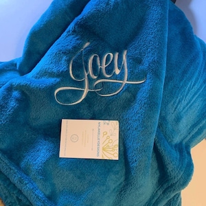 May include: A plush, teal blanket with the name "Joey" embroidered in white script. A business card from NYLAKELLEYDESIGNS rests on the blanket. The blanket is folded, showing its soft texture.