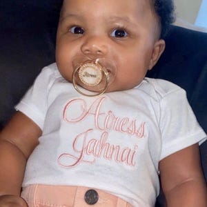 May include: A white short-sleeved bodysuit featuring the embroidered name Airess Jahnai in pink cursive script, paired with a gold-toned pacifier that has a circular center piece inscribed with the name Airess and a clear plastic shield.