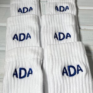 May include: White ribbed socks with the embroidered letters "ADA" in navy blue. The socks are folded and stacked, with a tag visible in the background. The text "NKDS" and "Thank You So Very Kind" is visible on the tag.