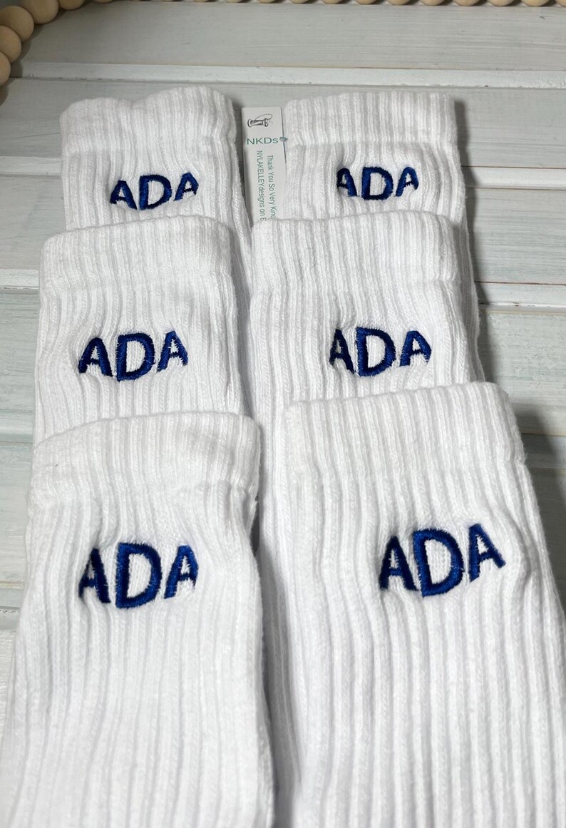 May include: White ribbed socks with the embroidered letters "ADA" in navy blue. The socks are folded and stacked, with a tag visible in the background. The text "NKDS" and "Thank You So Very Kind" is visible on the tag.