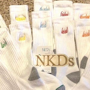 May include: A collection of white ribbed socks, each embroidered with a day of the week in various colors. The word "loved" is embroidered on each sock. A wooden sign with the text "NKDs" is in the center.