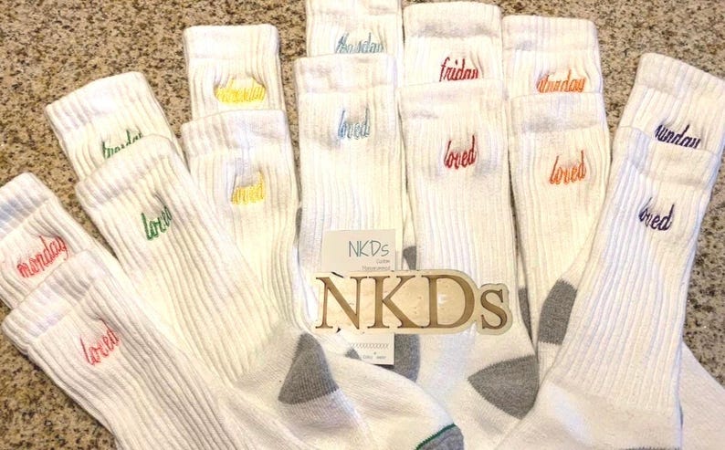 May include: A collection of white ribbed socks, each embroidered with a day of the week in various colors. The word "loved" is embroidered on each sock. A wooden sign with the text "NKDs" is in the center.