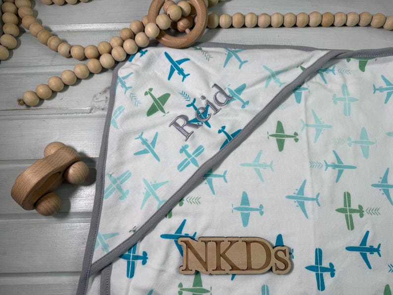 Puede incluir: Manta de beb&eacute; blanca con un estampado de aviones azules y verdes. La manta tiene un ribete gris y el nombre "Reid" bordado. Un avi&oacute;n de juguete de madera y una guirnalda de cuentas de madera tambi&eacute;n est&aacute;n en la imagen.
