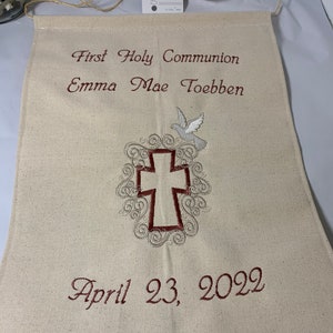 May include: A beige fabric banner embroidered with the words "First Holy Communion" and "Emma Mae Toebben" in burgundy thread. A cross and dove are embroidered below the text, with the date "April 23, 2022" at the bottom. A tag with the text "THANK YOU!" is attached.