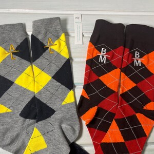 May include: Two pairs of argyle patterned socks. One pair is gray, yellow, and black with gold embroidered initials "A L". The other pair is red, orange, and brown with white embroidered initials "B M".
