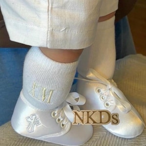 May include: White satin baby shoes with a cross design and ribbon laces. The shoes are paired with white socks embroidered with initials. The image also shows white linen pants. The text "NKDs" is visible on the shoes.