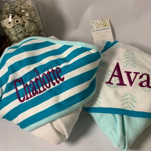 Puede incluir: Dos toallas con capucha para beb&eacute;. Una es blanca con rayas turquesas y blancas y el nombre "Charlotte" bordado en morado. La otra es blanca con un estampado de chevr&oacute;n turquesa y el nombre "Ava" bordado en morado. Las toallas tienen un ribete azul claro.