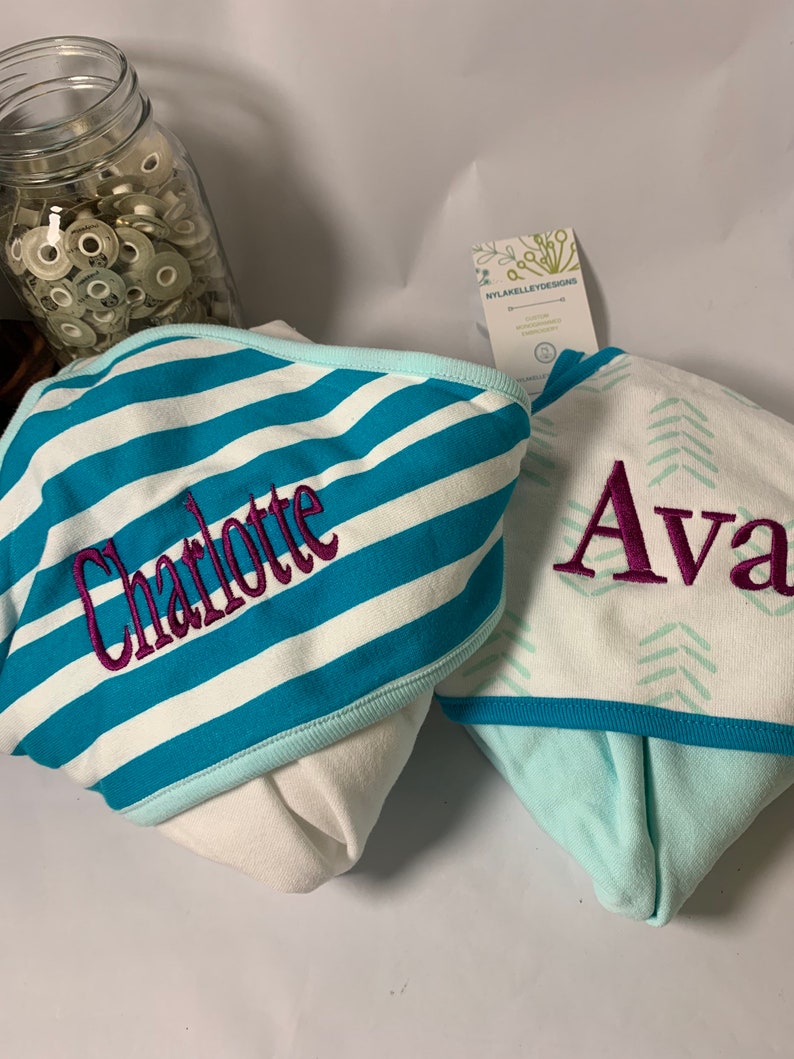 Puede incluir: Dos toallas con capucha para beb&eacute;. Una es blanca con rayas turquesas y blancas y el nombre "Charlotte" bordado en morado. La otra es blanca con un estampado de chevr&oacute;n turquesa y el nombre "Ava" bordado en morado. Las toallas tienen un ribete azul claro.