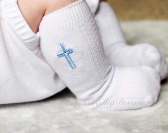 May include: White baby socks with a light blue embroidered cross design. The socks have a textured pattern and are designed for infants. The image shows a close-up of the socks, highlighting the cross detail.