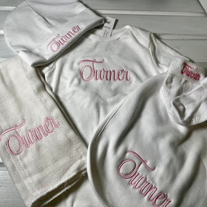 May include: A white baby set featuring a short-sleeve bodysuit, a beanie, a bib, and a pair of socks. Each item is embroidered with the name Turner in pink cursive script. The bodysuit, beanie, bib, and socks are made of white fabric.