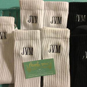 May include: White and black ribbed socks with embroidered initials. The white socks feature "JVM" or "JVN" in black thread, while the black socks have silver thread. A small card with text is also visible.