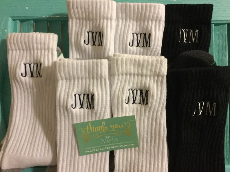 May include: White and black ribbed socks with embroidered initials. The white socks feature "JVM" or "JVN" in black thread, while the black socks have silver thread. A small card with text is also visible.