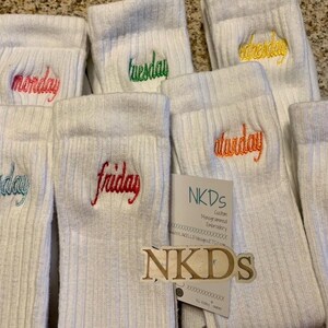 May include: White ribbed socks embroidered with the days of the week in various colors. The days are Monday, Tuesday, Wednesday, Thursday, Friday, Saturday, and Sunday. A tag with the text "NKDs" is also visible.