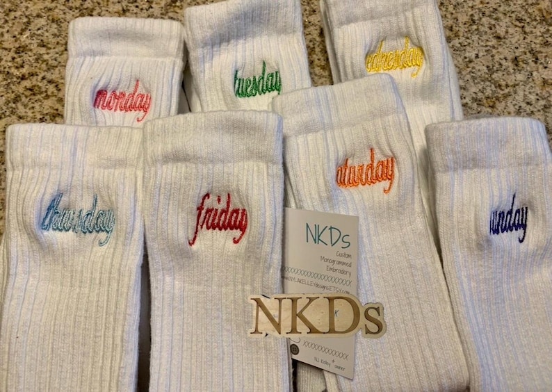 May include: White ribbed socks embroidered with the days of the week in various colors. The days are Monday, Tuesday, Wednesday, Thursday, Friday, Saturday, and Sunday. A tag with the text "NKDs" is also visible.