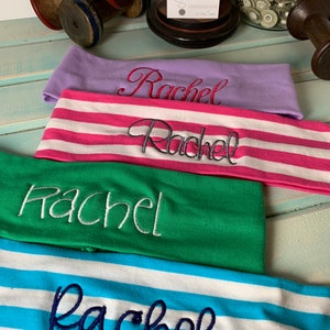 Pu&ograve; includere: Quattro fasce per capelli con il nome "Rachel" ricamato su di esse in diversi colori e font. Le fasce sono impilate l'una sull'altra. La fascia superiore &egrave; viola, la seconda &egrave; a righe rosa e bianche, la terza &egrave; verde e l'ultima &egrave; a righe blu e bianche.