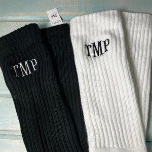 May include: Close-up of two pairs of ribbed knit socks. One pair is black, the other is white. Each sock has the embroidered letters "TMP" in the same color as the sock. The socks are displayed on a light blue surface. A tag with the text "NKDs" is visible.