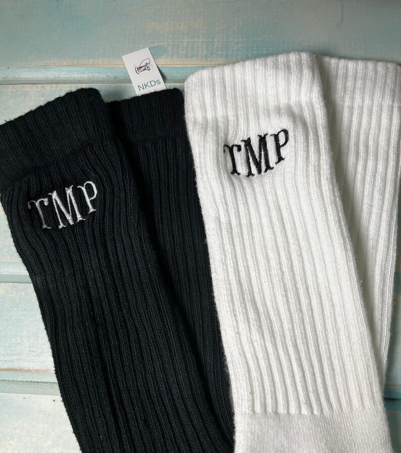 May include: Close-up of two pairs of ribbed knit socks. One pair is black, the other is white. Each sock has the embroidered letters "TMP" in the same color as the sock. The socks are displayed on a light blue surface. A tag with the text "NKDs" is visible.