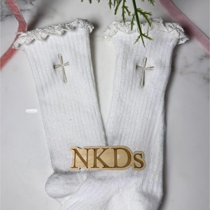 May include: A pair of white socks with a delicate lace trim at the top and an embroidered cross near the ankle. The socks have a ribbed texture and a non-slip grip on the sole. A wooden sign with the letters "NKDs" is also present.