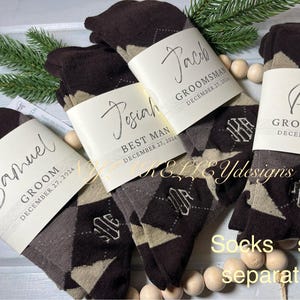 May include: Four pairs of brown and beige argyle socks with personalized gift tags. The tags are cream colored with black calligraphy and include the names Samuel, Josiah, Jack, and Paul. The tags also include the titles Groom, Best Man, and Groomsmans, and the date December 27, 2024. The text "Socks sold separately" is printed at the bottom of the image.