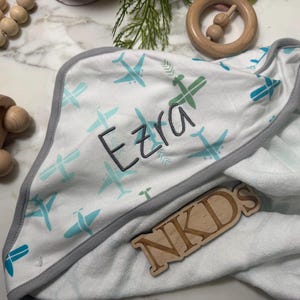 Puede incluir: Toalla con capucha blanca para beb&eacute; con ribete gris y estampado de aviones en verde y turquesa. El nombre "Ezra" est&aacute; bordado en la toalla. Tambi&eacute;n se ven juguetes de madera, como un coche y un mordedor.