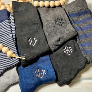 May include: An assortment of socks in various colors and patterns. The socks include blue and gray striped, solid black, gray, and blue socks. Some socks feature embroidered initials in white or gold thread. A wooden bead garland is also present.