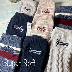 May include: A set of six pairs of cozy knit socks with personalized names embroidered on them. The socks are in various colors, including gray, white, blue, and burgundy. The text "Super Soft & Cozy" is written on the bottom of the image.