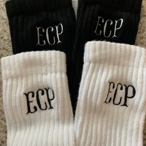 May include: A pair of black and a pair of white ribbed socks. Each sock has the embroidered letters "ECP" in a contrasting color. The socks are folded over, showing the ribbed cuff. The background is a neutral color.