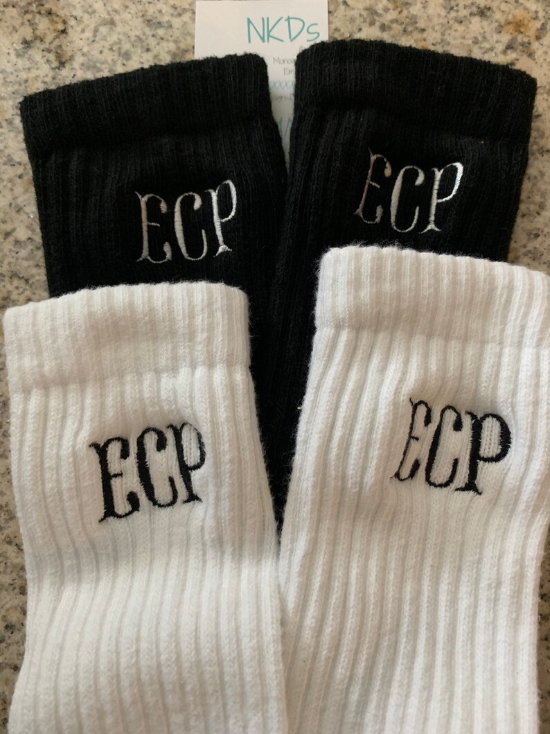 May include: A pair of black and a pair of white ribbed socks. Each sock has the embroidered letters "ECP" in a contrasting color. The socks are folded over, showing the ribbed cuff. The background is a neutral color.