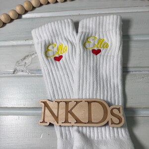 May include: White ribbed socks with the name "Ella" embroidered in yellow script above a red heart. A wooden sign with the letters "NKDs" is placed on the socks. The socks have gray heels and toes.