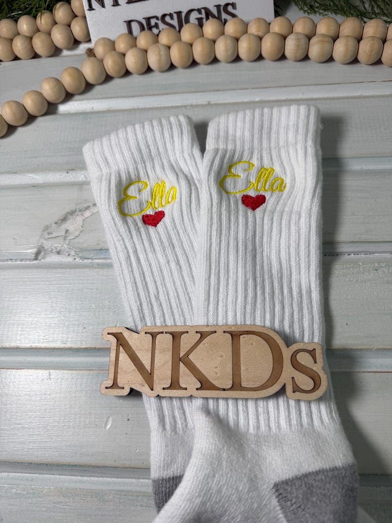 May include: White ribbed socks with the name "Ella" embroidered in yellow script above a red heart. A wooden sign with the letters "NKDs" is placed on the socks. The socks have gray heels and toes.