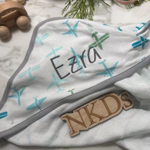 Puede incluir: Toalla con capucha para beb&eacute; blanca con ribete gris y un estampado de aviones azules y verdes. El nombre "Ezra" est&aacute; bordado en gris. Tambi&eacute;n hay un letrero de madera con las letras "NKDS".