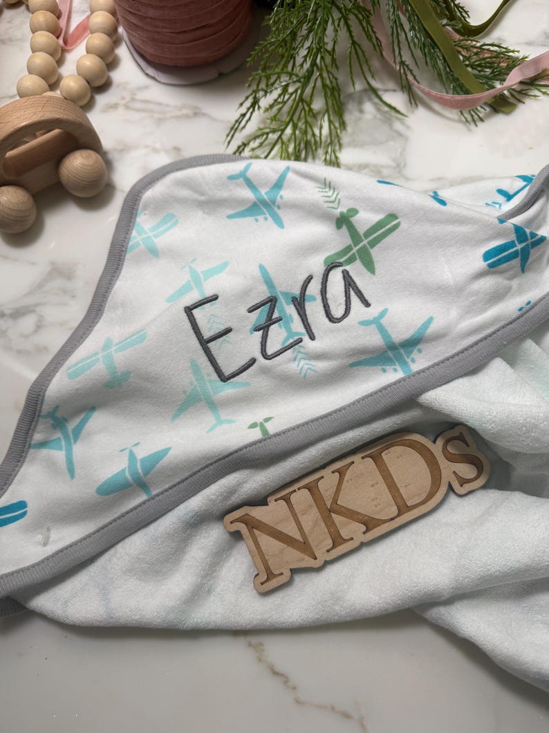 Puede incluir: Toalla con capucha para beb&eacute; blanca con ribete gris y un estampado de aviones azules y verdes. El nombre "Ezra" est&aacute; bordado en gris. Tambi&eacute;n hay un letrero de madera con las letras "NKDS".