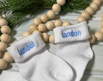 以下が含まれることがあります： 青い糸でLandonという名前が刺繍されたリブ編みのカフが付いた白い乳幼児用靴下。靴下は、NYLAKELLEYDESIGNSという文字が書かれた白い名刺と、天然木のビーズの紐と一緒に配置されています。