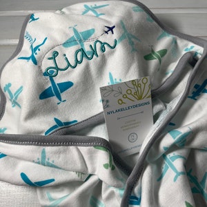 Puede incluir: Manta de beb&eacute; blanca con un estampado de aviones turquesa y azul claro. La manta tiene un ribete gris y el nombre "Liam" est&aacute; bordado en hilo turquesa. Una etiqueta de "NYLAKELLEYDESIGNS" est&aacute; adjunta.