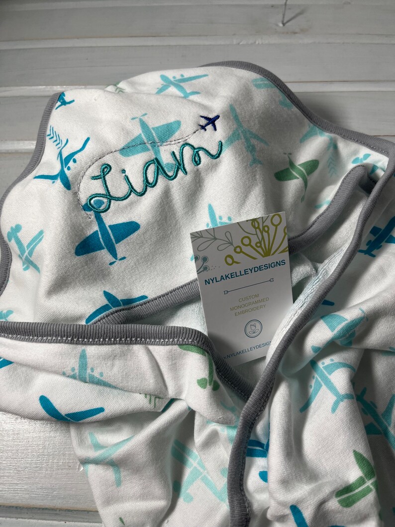 Puede incluir: Manta de beb&eacute; blanca con un estampado de aviones turquesa y azul claro. La manta tiene un ribete gris y el nombre "Liam" est&aacute; bordado en hilo turquesa. Una etiqueta de "NYLAKELLEYDESIGNS" est&aacute; adjunta.