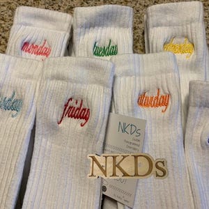 May include: White ribbed socks embroidered with the days of the week in various colors. The days are Monday, Tuesday, Wednesday, Thursday, Friday, Saturday, and Sunday. A tag with the brand name "NKDs" is also visible.