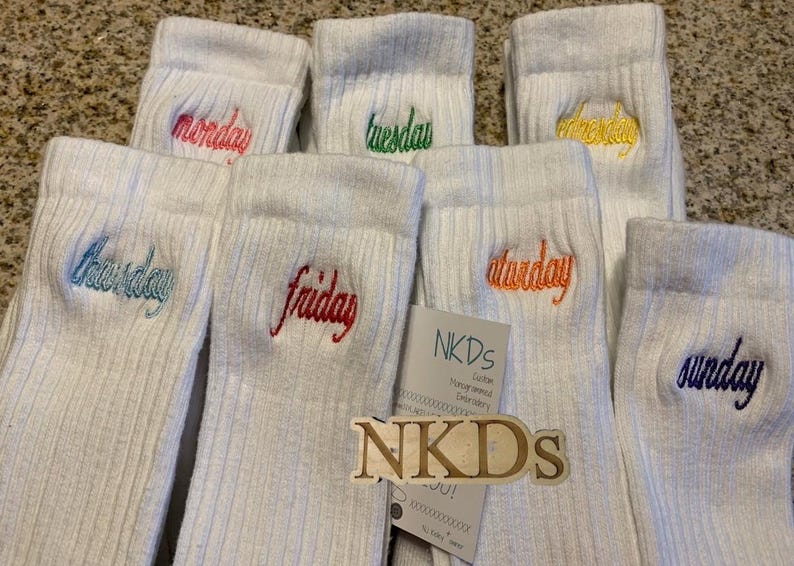 May include: White ribbed socks embroidered with the days of the week in various colors. The days are Monday, Tuesday, Wednesday, Thursday, Friday, Saturday, and Sunday. A tag with the brand name "NKDs" is also visible.