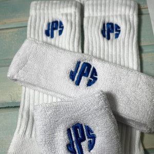 May include: A set of white athletic accessories including two ribbed crew socks, a headband, and a wristband. Each item features the embroidered blue monogram letters JPS in a circular style. The socks have grey toe and heel sections.