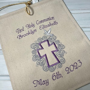 May include: A white canvas banner with purple embroidery. The banner reads "First Holy Communion Brooklyn Elizabeth May 6th, 2023". A purple cross with a white dove is embroidered in the center.