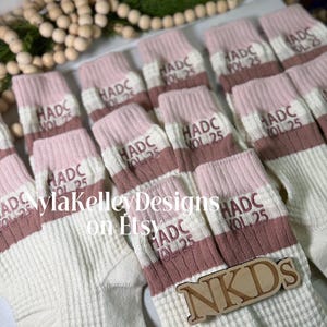 May include: A collection of cream-colored socks with pink and brown stripes. Each sock features the text "HADC VOL 25" in brown. A wooden sign with "NKDS" is placed on top of the socks. The Etsy shop name "NylakelleyDesigns" is also visible.