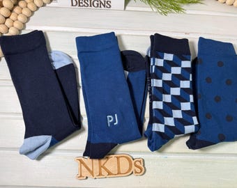Può includere: Quattro paia di calze in varie tonalità di blu. Un paio è blu navy tinta unita con talloni e punte azzurri, uno è blu medio con le lettere ricamate PJ, uno presenta un motivo geometrico a cubi e uno ha una stampa a pois blu scuro.